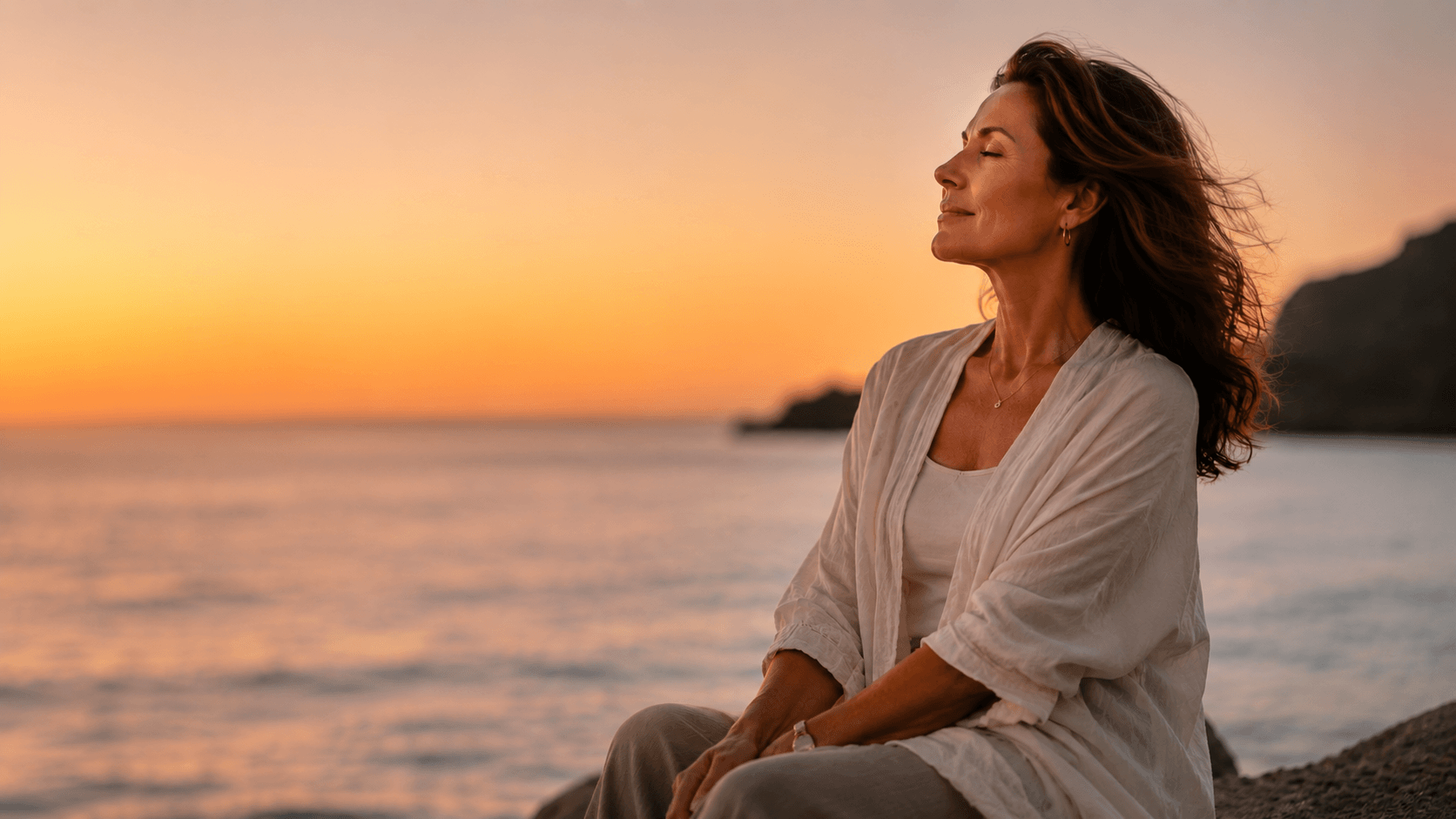 Soft peaceful woman sitting on a beach with her hair blowing in the wind during sunset